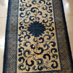 Low-pile tan rug featuring a central dark blue floral design and a geometric fret border on light tile.