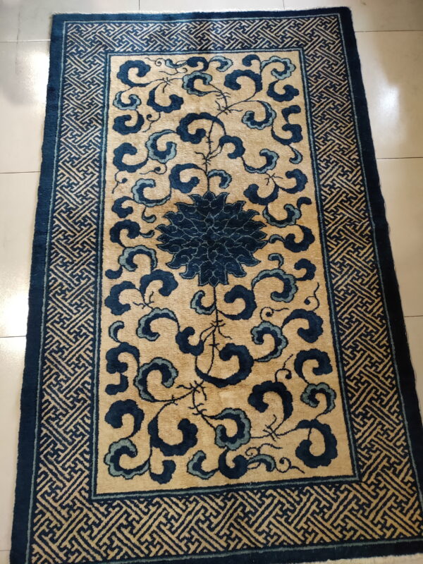 Low-pile tan rug featuring a central dark blue floral design and a geometric fret border on light tile.