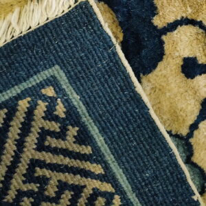 Close-up of a dark blue and tan high-pile rug showing the woven edge, geometric border, and fringe.