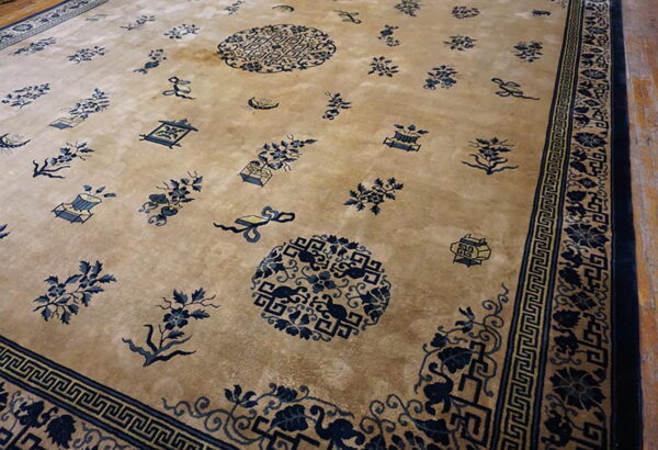 Angled view of a tan rug with scattered blue oriental motifs and geometric banding along the border.