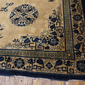 Golden tan rug with a navy blue border showing Greek key and floral patterns on worn wood flooring.