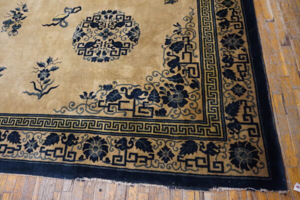 Golden tan rug with a navy blue border showing greek key and floral patterns on worn wood flooring.