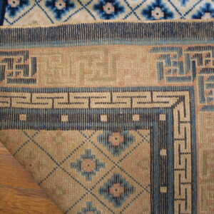 The corner of a low-pile tan and navy geometric rug is folded over a white and blue patterned rug on wood floor.