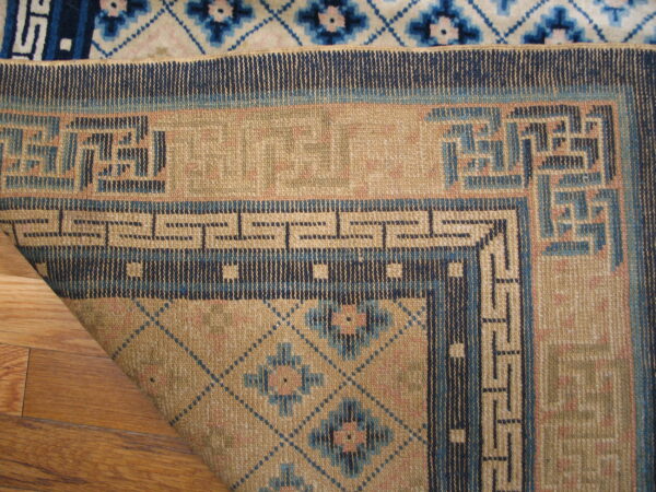 The corner of a low-pile tan and navy geometric rug is folded over a white and blue patterned rug on wood floor.