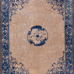 Tan area rug with a dark blue traditional border and medallion lies on worn wooden flooring.