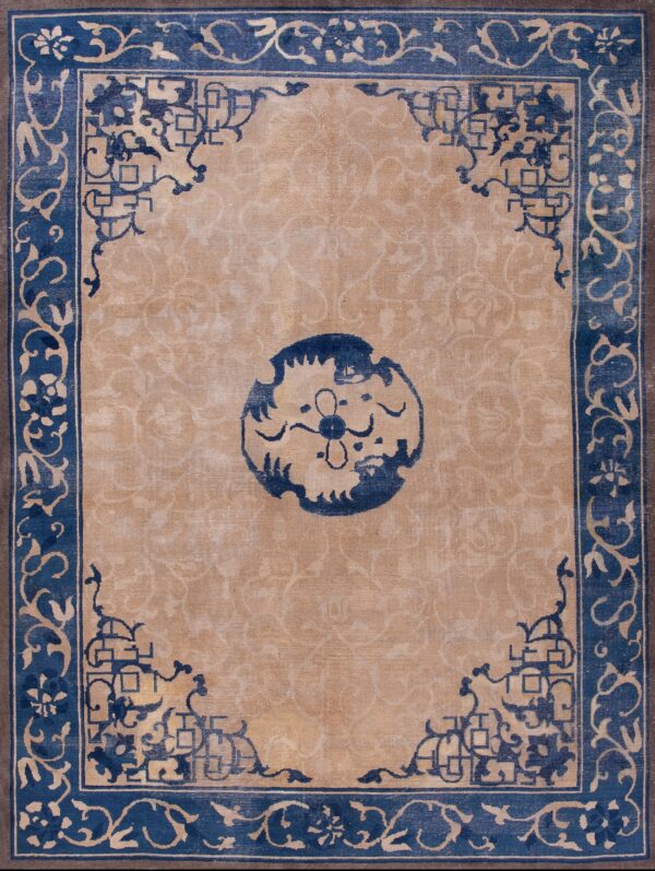 Tan area rug with a dark blue traditional border and medallion lies on worn wooden flooring.