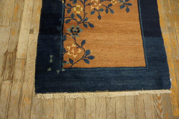 Navy border frames a tan rug with blue and cream floral vines on rustic plank wood flooring.