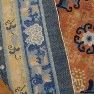 Close-up of a folded rug showing deep orange pile beside a tightly woven blue and yellow striped border on wood floor.