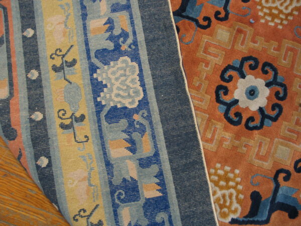 Close-up of a folded rug showing deep orange pile beside a tightly woven blue and yellow striped border on wood floor.