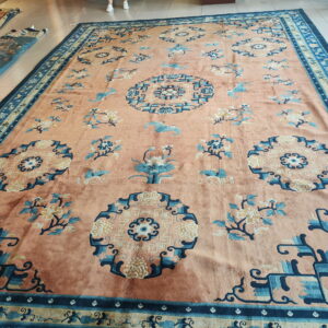 Low-pile salmon rug with blue geometric corner medallions and scattered floral motifs on white tile flooring.
