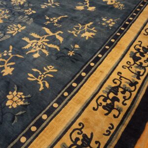 Deep blue rug with gold botanical patterns and wide scrolling borders lies on light brown wood flooring.
