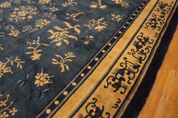 Deep blue rug with gold botanical patterns and wide scrolling borders lies on light brown wood flooring.