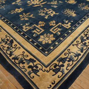 Blue and tan low-pile rug featuring stylized botanical patterns and a geometric corner border resting on wood planks.