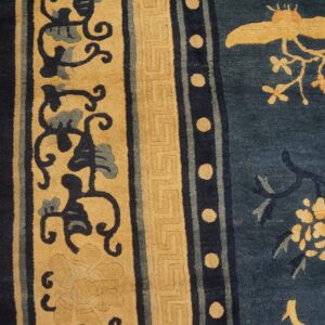 Indigo and tan rug displays detailed borders, scrolling vines, and a central bird motif over wood planks.