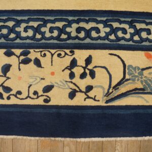 Beige rug with dark blue scroll and floral patterns, visible fringe, resting on wood flooring.