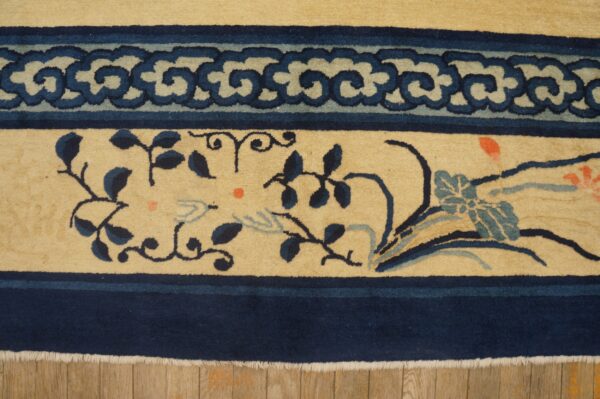Beige rug with dark blue scroll and floral patterns, visible fringe, resting on wood flooring.