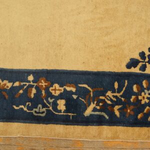 High-pile tan rug with a navy floral border and corner detail rests on light hardwood flooring.