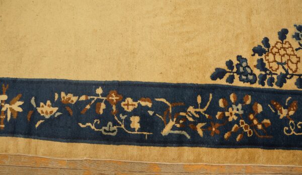 High-pile tan rug with a navy floral border and corner detail rests on light hardwood flooring.