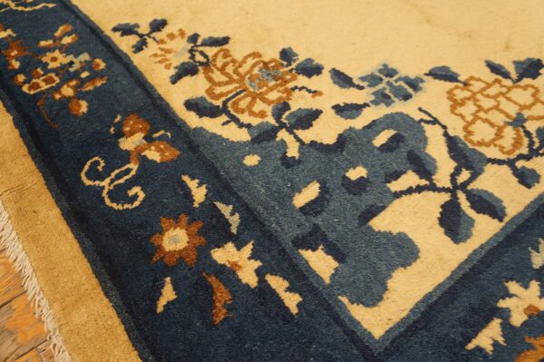 Low-pile rug in tan and navy blue with floral patterns, resting on visible wooden flooring.