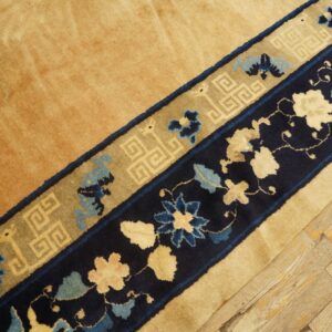 Close-up of a gold rug with navy blue and tan decorative borders on distressed wood flooring.