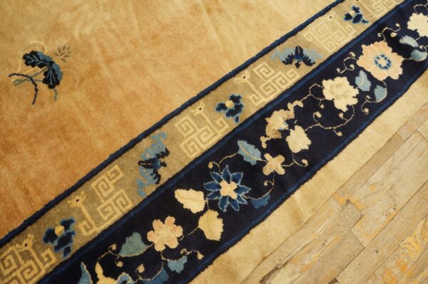 Close-up of a gold rug with navy blue and tan decorative borders on distressed wood flooring.