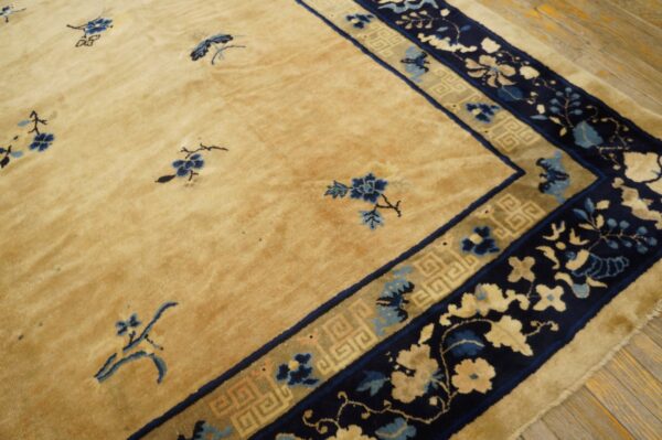 Tan area rug featuring sparse blue florals and an ornate navy and gold multi-layered border on wood flooring.