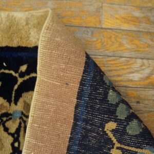 A rolled rug shows its beige woven backing and navy and tan floral high pile on distressed wood flooring.