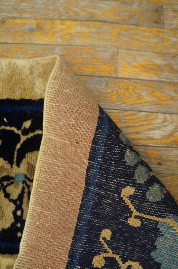 A rolled rug shows its beige woven backing and navy and tan floral high pile on distressed wood flooring.