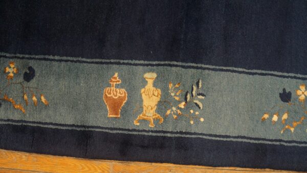 Dark blue rug with a slate border showing ornate tan and rust vases and foliage on a wood floor.