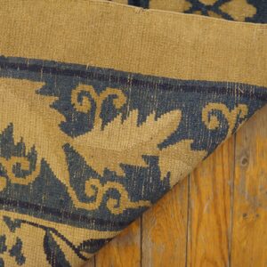 Low-pile blue and tan rug showing a detailed foliage pattern, folded over rustic amber wood planks.