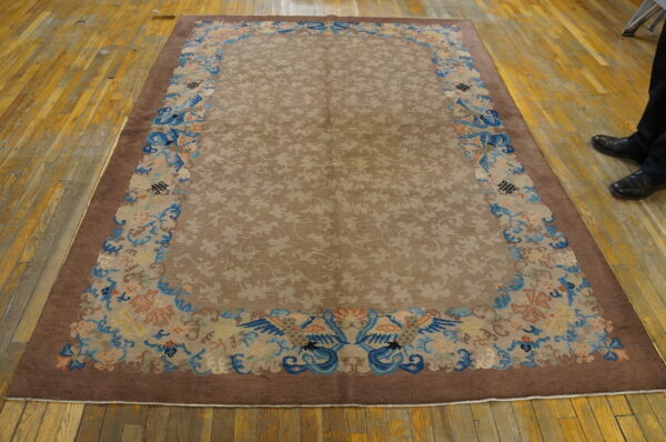 A brown rug with ornate blue and peach bird borders lies on a worn wooden floor beside black shoes.