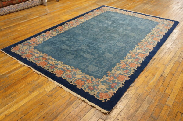 Low-pile rug in dusty blue with a floral center and decorative coral border on wood floors.