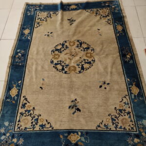 Low-pile rug in beige and deep blue featuring a floral border and central medallion on white tile flooring.