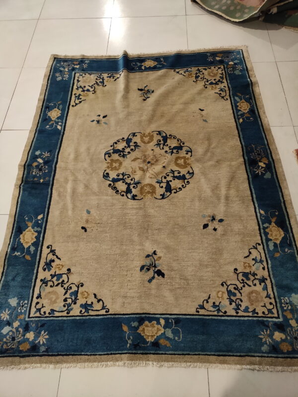 Low-pile rug in beige and deep blue featuring a floral border and central medallion on white tile flooring.