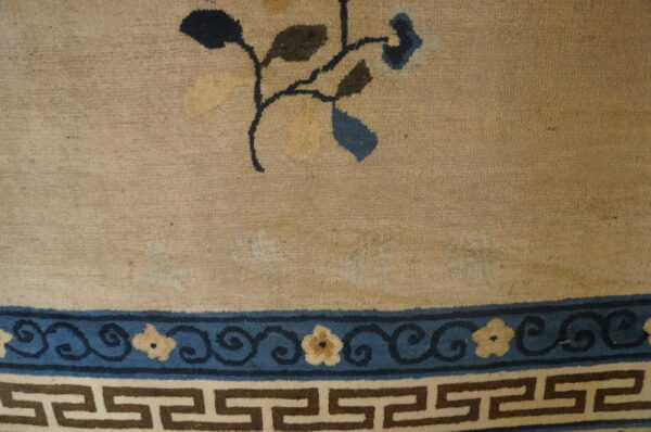 Close-up of a tan rug field with dark floral elements above a complex blue and brown geometric border.