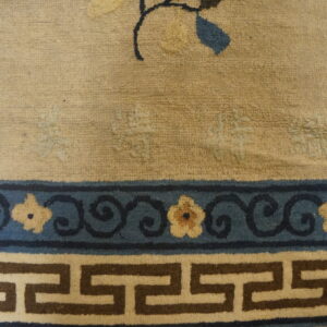 Beige textured rug features a border with dark blue floral scrollwork above a brown and cream Greek key band.