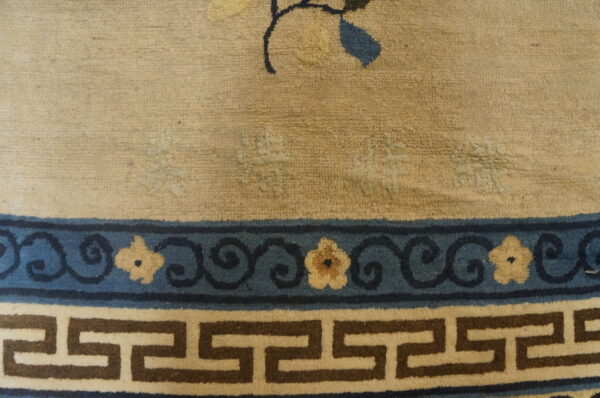 Beige textured rug features a border with dark blue floral scrollwork above a brown and cream greek key band.