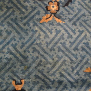 Muted blue rug with an interlocking geometric pattern, tan floral details, and a black and tan border.