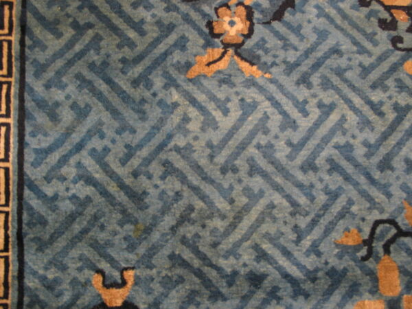 Muted blue rug with an interlocking geometric pattern, tan floral details, and a black and tan border.