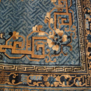 Corner detail of a blue and tan rug with stylized floral and geometric fretwork borders on wood flooring.