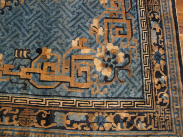 Corner detail of a blue and tan rug with stylized floral and geometric fretwork borders on wood flooring.