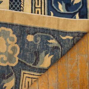 Blue and beige patterned rug folded over, displaying its backing on distressed wood planks.
