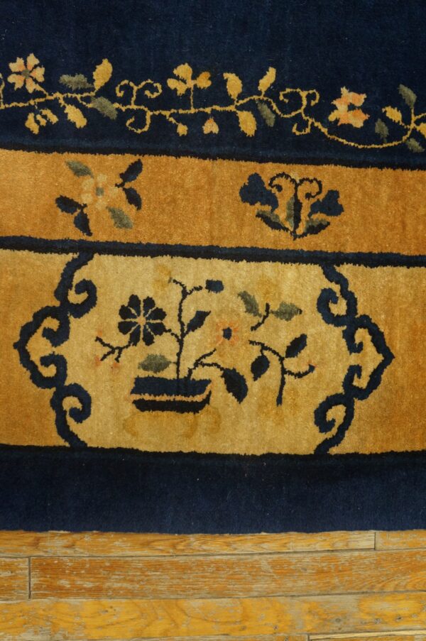 Blue antique rectangle 1920s wool hand knotted pile rug 6 Detail of a navy and gold high-pile rug featuring a stylized potted plant motif over distressed wood flooring.