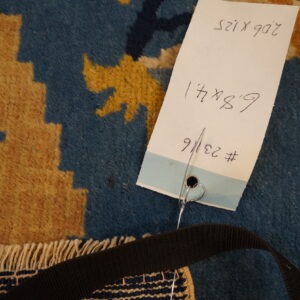 Blue and golden patterned rug detail with dense pile, white fringe, and an attached inventory tag.