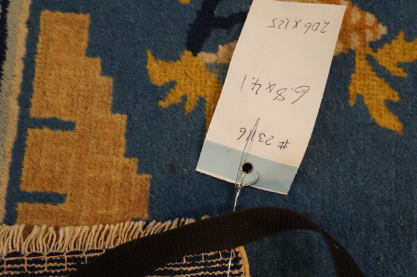 Blue and golden patterned rug detail with dense pile, white fringe, and an attached inventory tag.