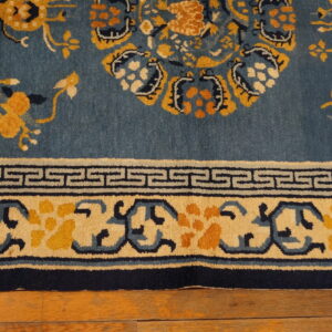 A low-pile rug featuring blue, yellow, and navy floral designs and a Greek key border over hardwood floorboards.