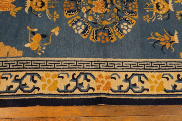 A low-pile rug featuring blue, yellow, and navy floral designs and a greek key border over hardwood floorboards.