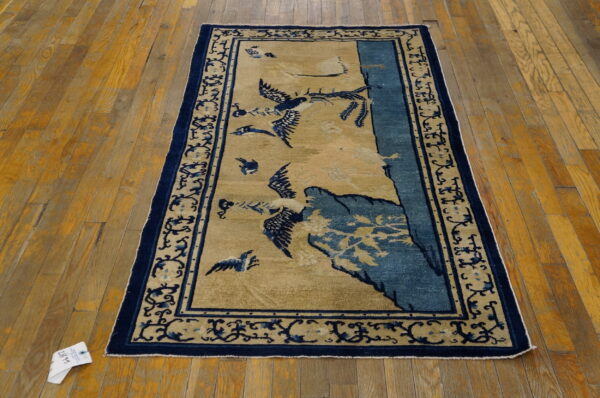 Low-pile tan and navy rug featuring flying bird and mountain designs, bordered by scrolling vines, lying on wood floors.