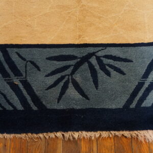 High pile tan rug edge with a dark blue bamboo patterned border and cream fringe on wood flooring.