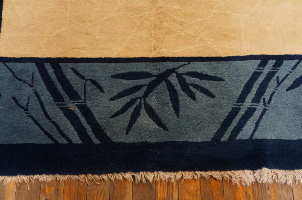 High pile tan rug edge with a dark blue bamboo patterned border and cream fringe on wood flooring.